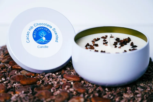 Candle with cacao nibs in a white container on a surface with cacao nibs.