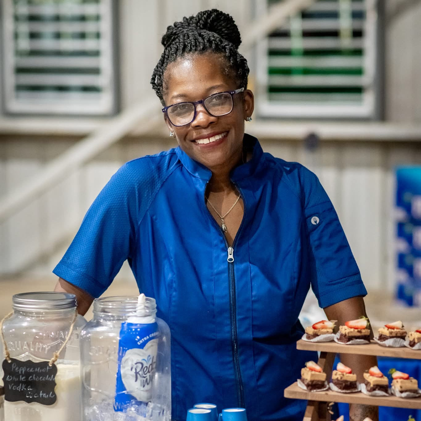 Erika Dupree Cline in a blue outfit standing behind a table with various items including jars, cups, and a display of cupcakes.