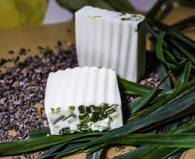 Two blocks of white chocolate soap with green lemongrass on a bed of purple stones and green leaves.